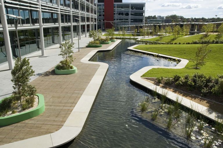 Surrounded by six large floor plate office buildings, the internal courtyard forms the focus for the Optus corporate campus in North Ryde. A 200 metre long ribbon wall unifies the space and articulates the straight podium edge to create a number of sub spaces within the triangular courtyard. Stairs and planted embankments rise up to the ribbon wall to take up the four metre level change between the podium and the courtyard to its south. Storm water detention is handled in two large ponds with an elegant precast edge. Planting is scaled according to the spaces. Tall trees are planted on the embankments and small ornamental tress within the sub spaces at the base of the ribbon wall. The project sought to provide the employees with a variety of richly planted garden spaces in which to gather, socialise and hang out during their breaks. The spatial structure is reinforced by the planting palette. Tall straight trunked spotted gums line the ramps and signify main circulation spaces, whilst small flowering deciduous trees and flowering shrubs define the sub spaces. The ponds are planted with water plants.