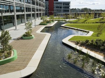 Surrounded by six large floor plate office buildings, the internal courtyard forms the focus for the Optus corporate campus in North Ryde. A 200 metre long ribbon wall unifies the space and articulates the straight podium edge to create a number of sub spaces within the triangular courtyard. Stairs and planted embankments rise up to the ribbon wall to take up the four metre level change between the podium and the courtyard to its south. Storm water detention is handled in two large ponds with an elegant precast edge. Planting is scaled according to the spaces. Tall trees are planted on the embankments and small ornamental tress within the sub spaces at the base of the ribbon wall. The project sought to provide the employees with a variety of richly planted garden spaces in which to gather, socialise and hang out during their breaks. The spatial structure is reinforced by the planting palette. Tall straight trunked spotted gums line the ramps and signify main circulation spaces, whilst small flowering deciduous trees and flowering shrubs define the sub spaces. The ponds are planted with water plants.